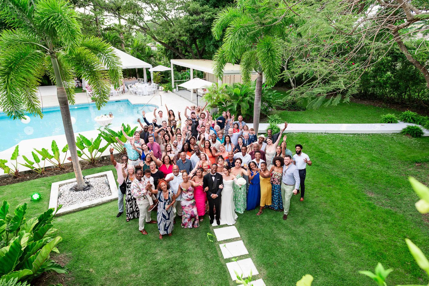 Large wedding group photo with guests gathered on green lawn near swimming pool.