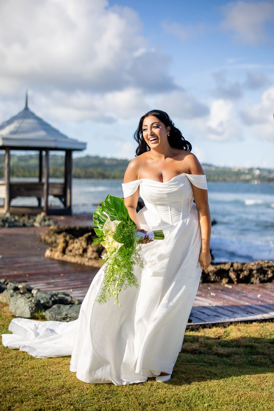 Elegant white wedding dress photographed at oceanfront venue with gazebo and grassy lawn.