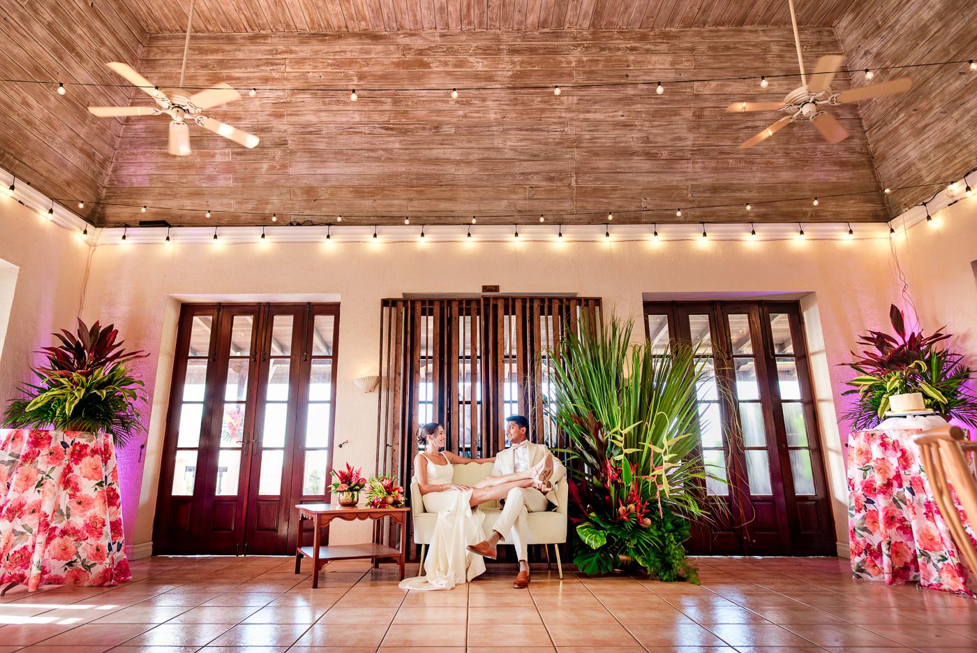 Elegant wedding reception venue with string lights, French doors, and tropical floral arrangements.