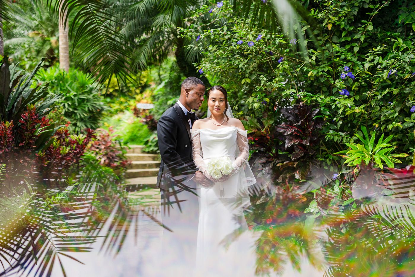 Wedding couple embraces in lush tropical garden setting with palm fronds.