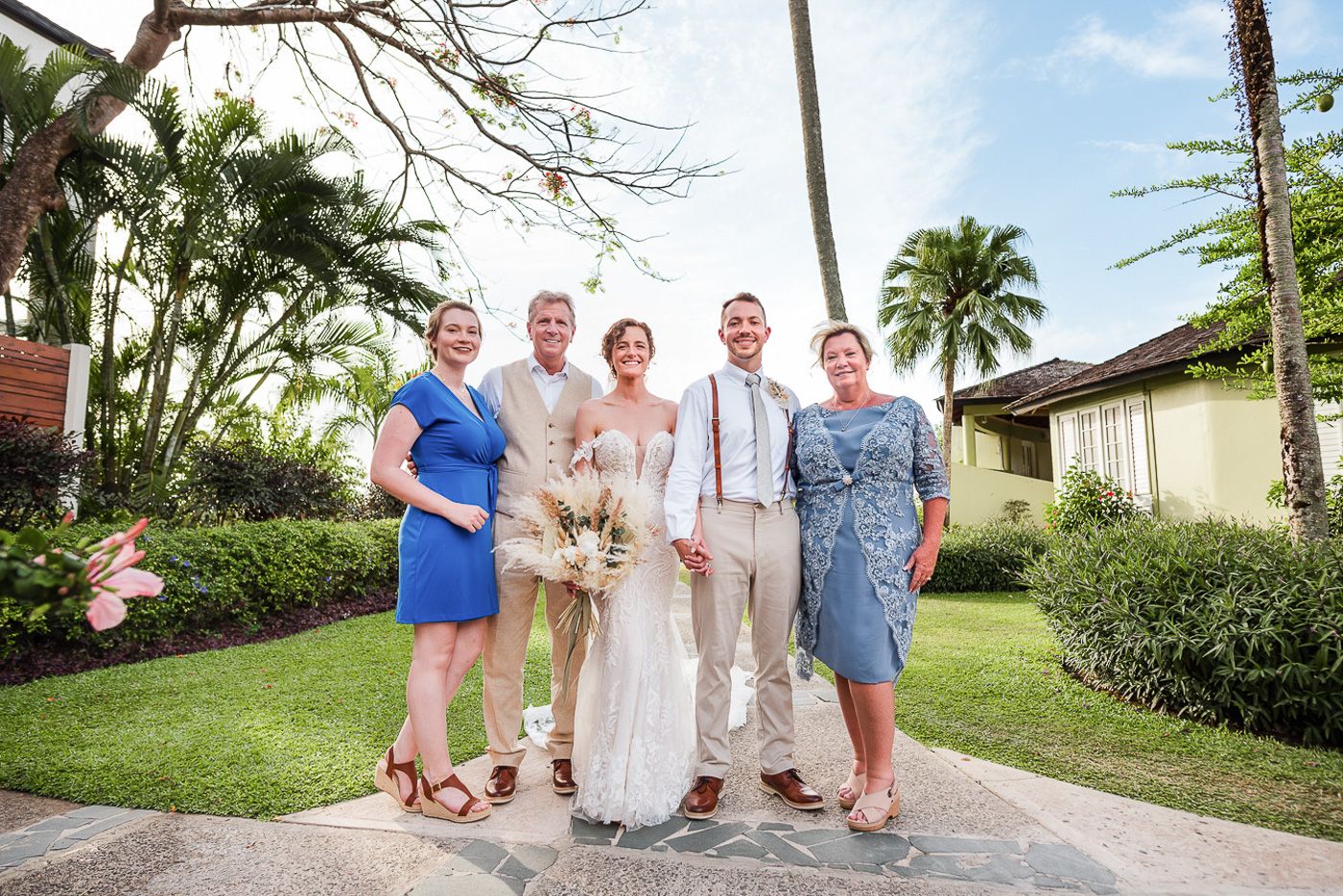 Wedding party members gather outdoors under palm trees in tropical setting.