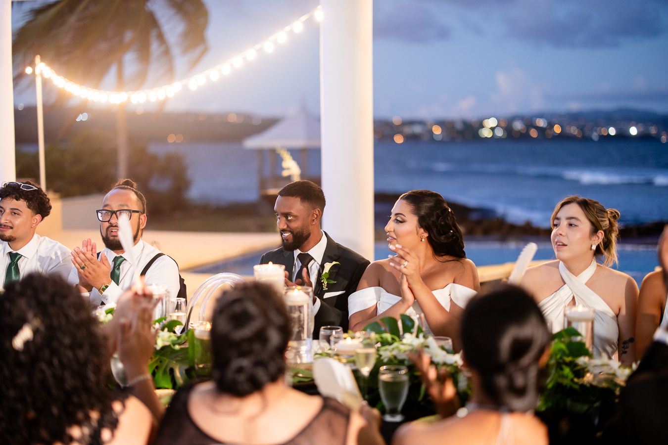 Wedding reception guests enjoying celebration under string lights by the ocean.