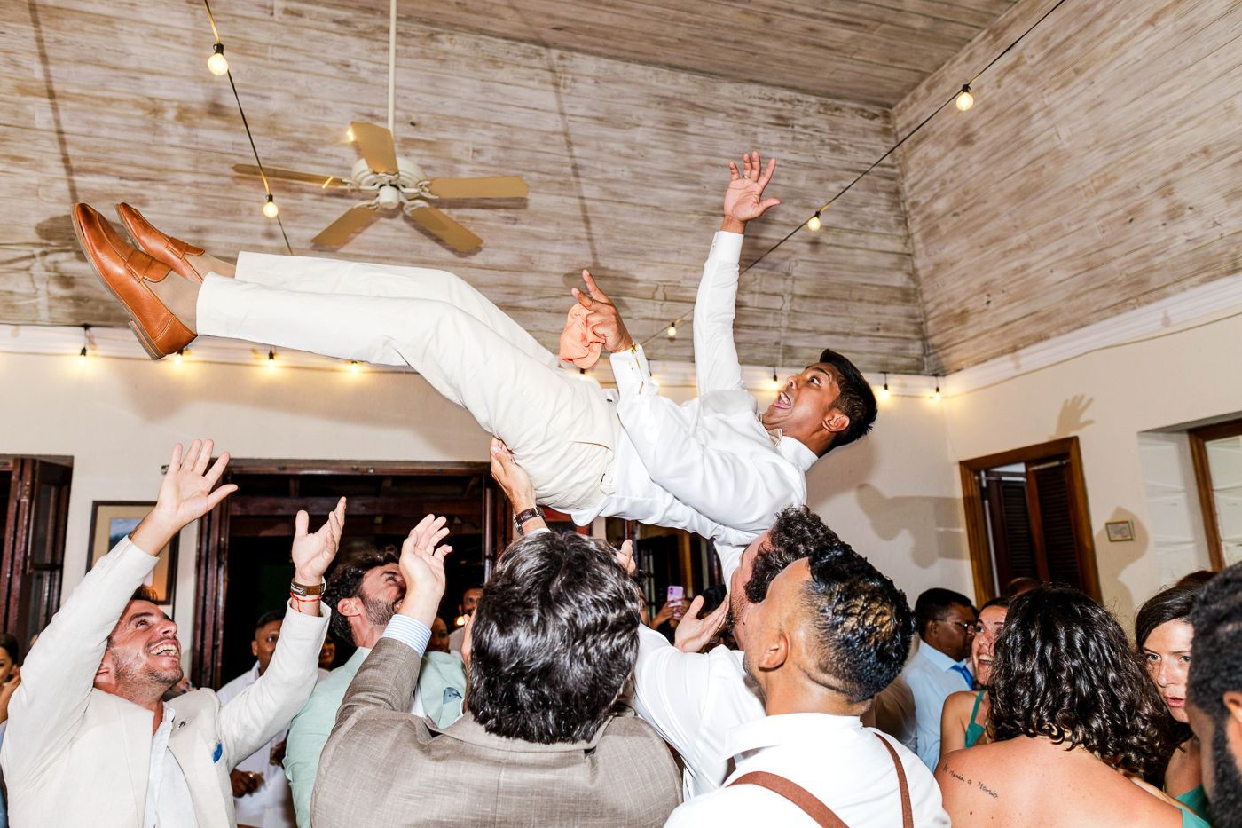 People dancing and raising their hands under string lights on a rustic wooden ceiling.