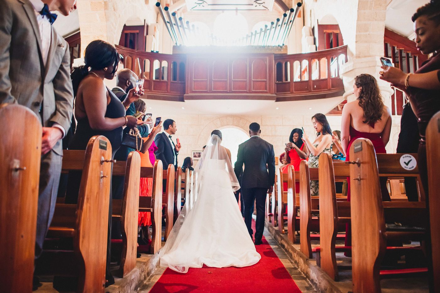 Wedding procession down a red-carpeted church aisle with wooden pews filled with guests.