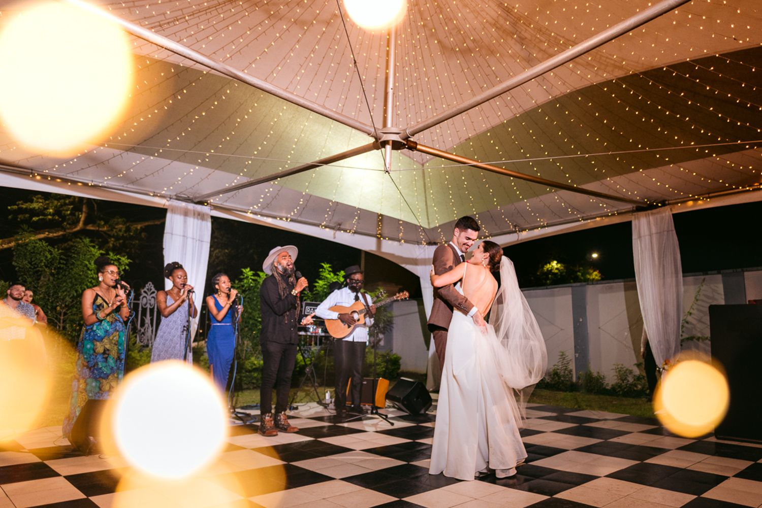 A wedding reception with string lights overhead and couples dancing on a black and white checkered dance floor.