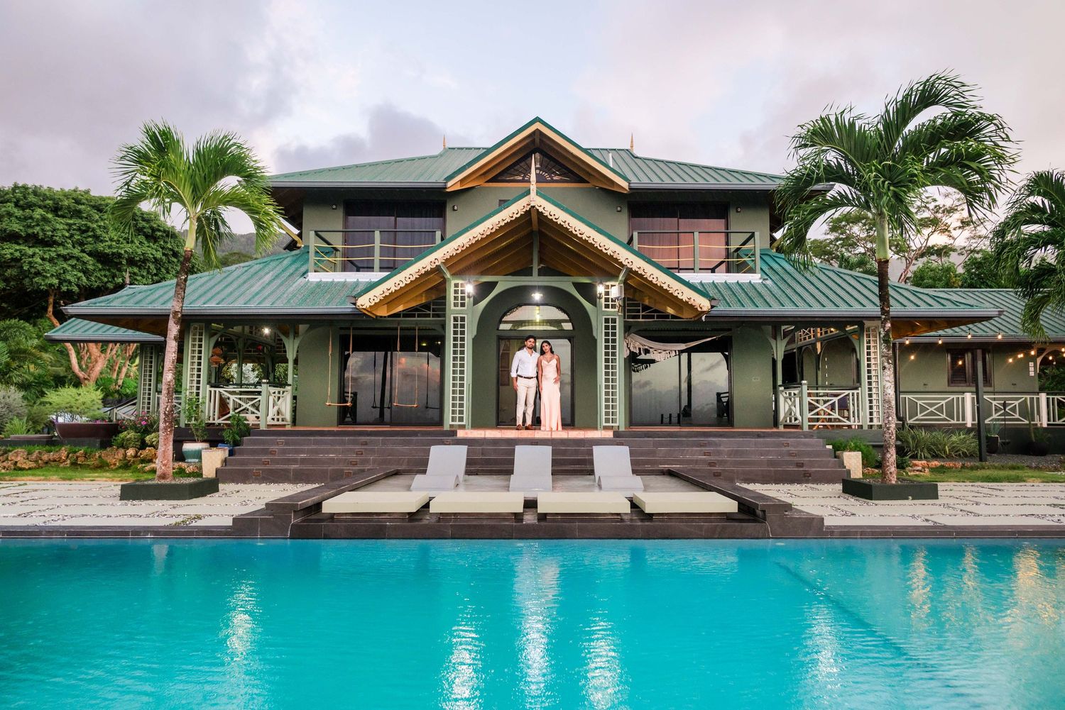 Couple standing on steps of luxury tropical villa with green roof, palm trees, and turquoise swimming pool.