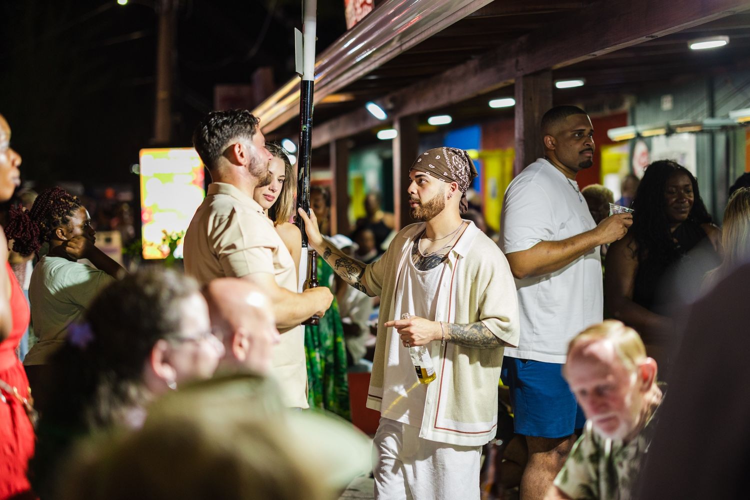 Crowd of people socializing outside a busy street venue at night, with a man in a white outfit and bandana.
