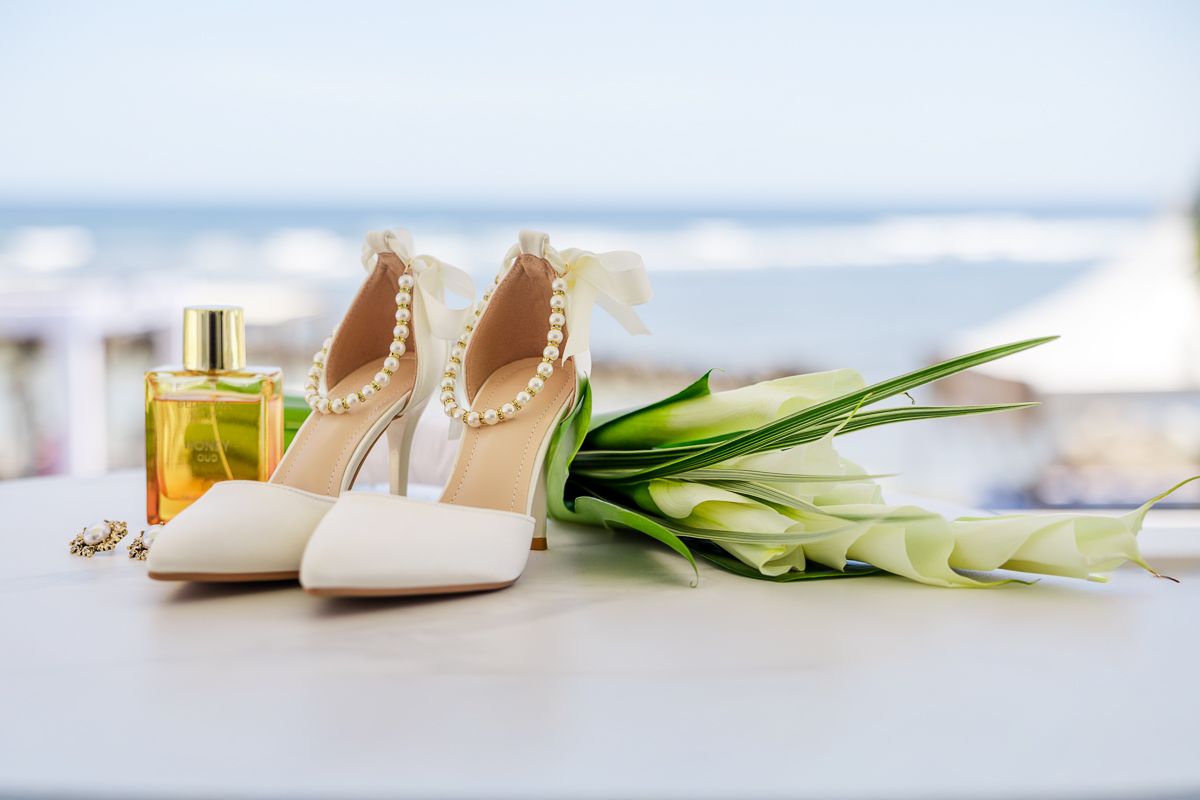 White pearl-adorned bridal heels beside a gold perfume bottle and white calla lilies on a table with ocean backdrop.