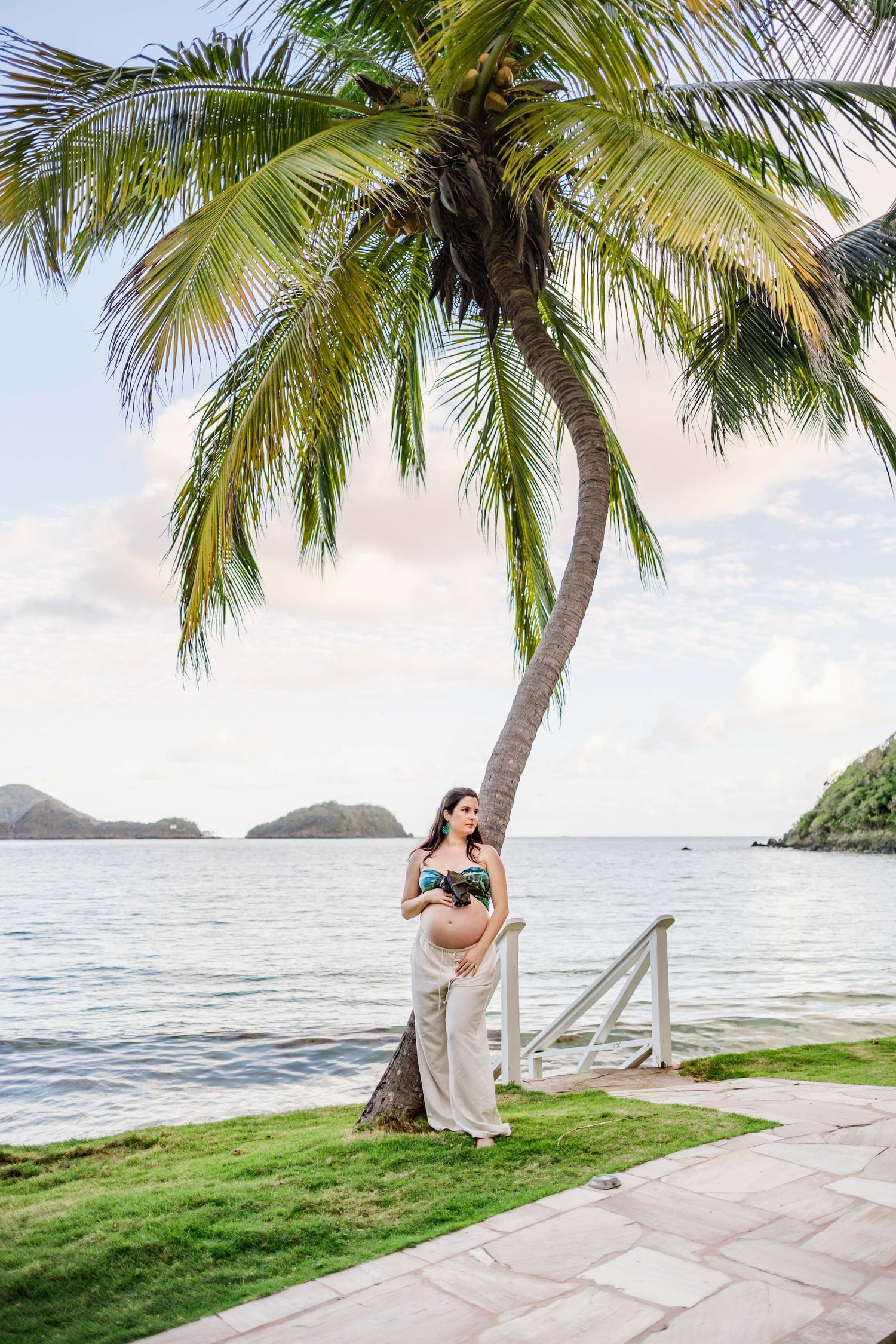 Pregnant woman in bikini top and white pants standing under a palm tree by the ocean at a tropical resort.