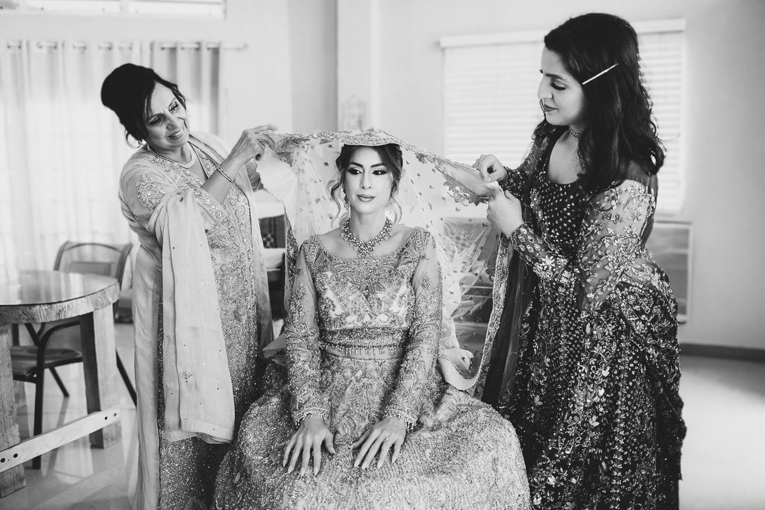 Black and white photo of a bride in ornate gown being helped with her crown by two women in traditional attire.