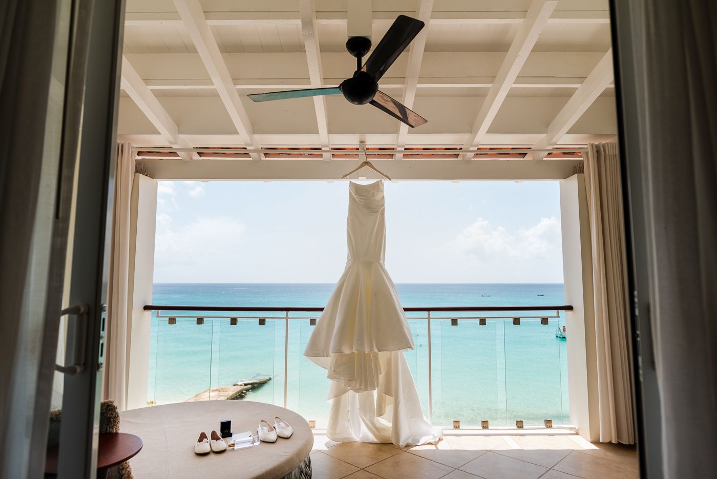 Bridal gown hangs in window frame with ocean view and natural lighting.