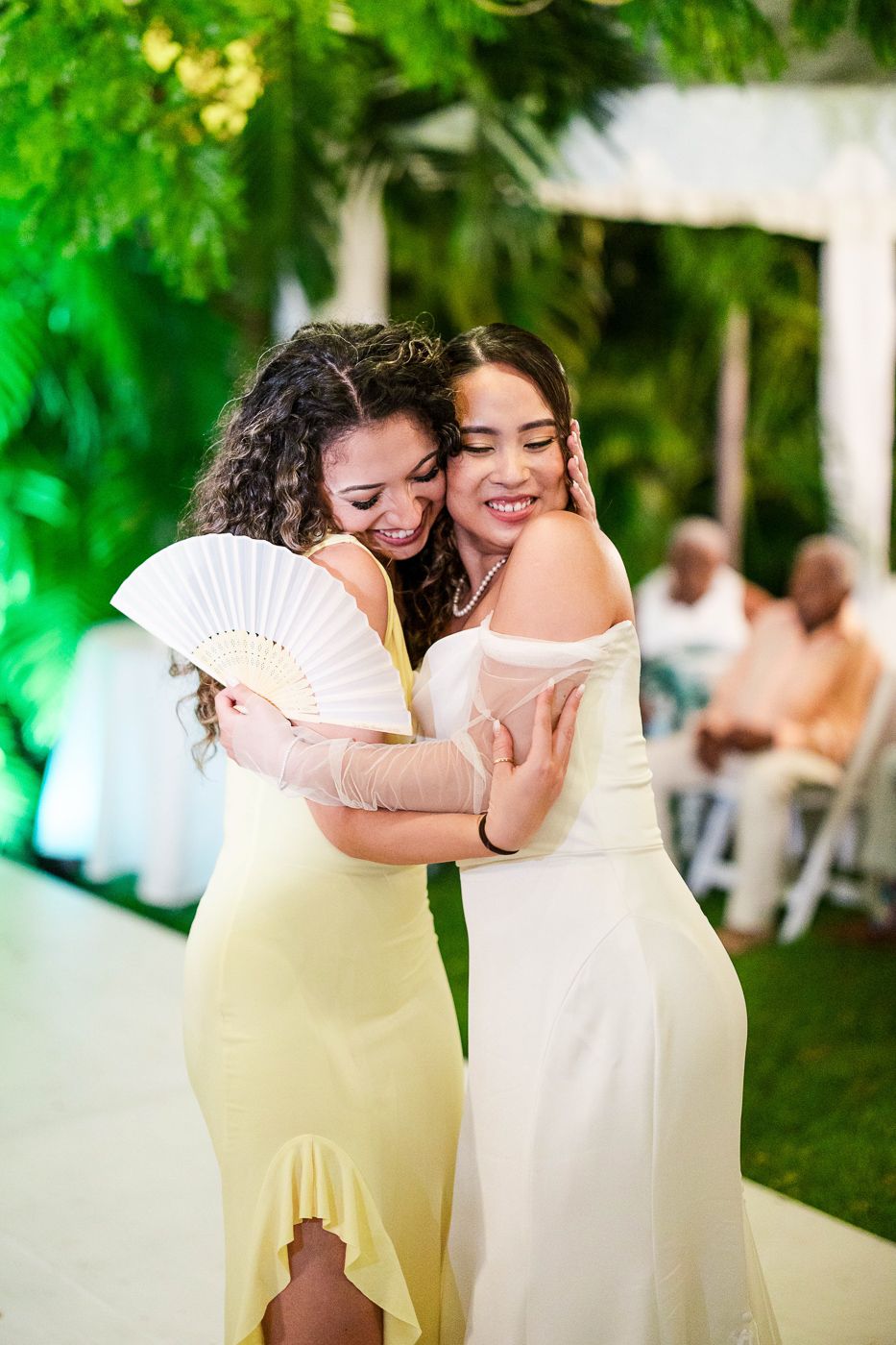 Two people in pastel dresses share an intimate moment while holding decorative fans at an outdoor celebration.