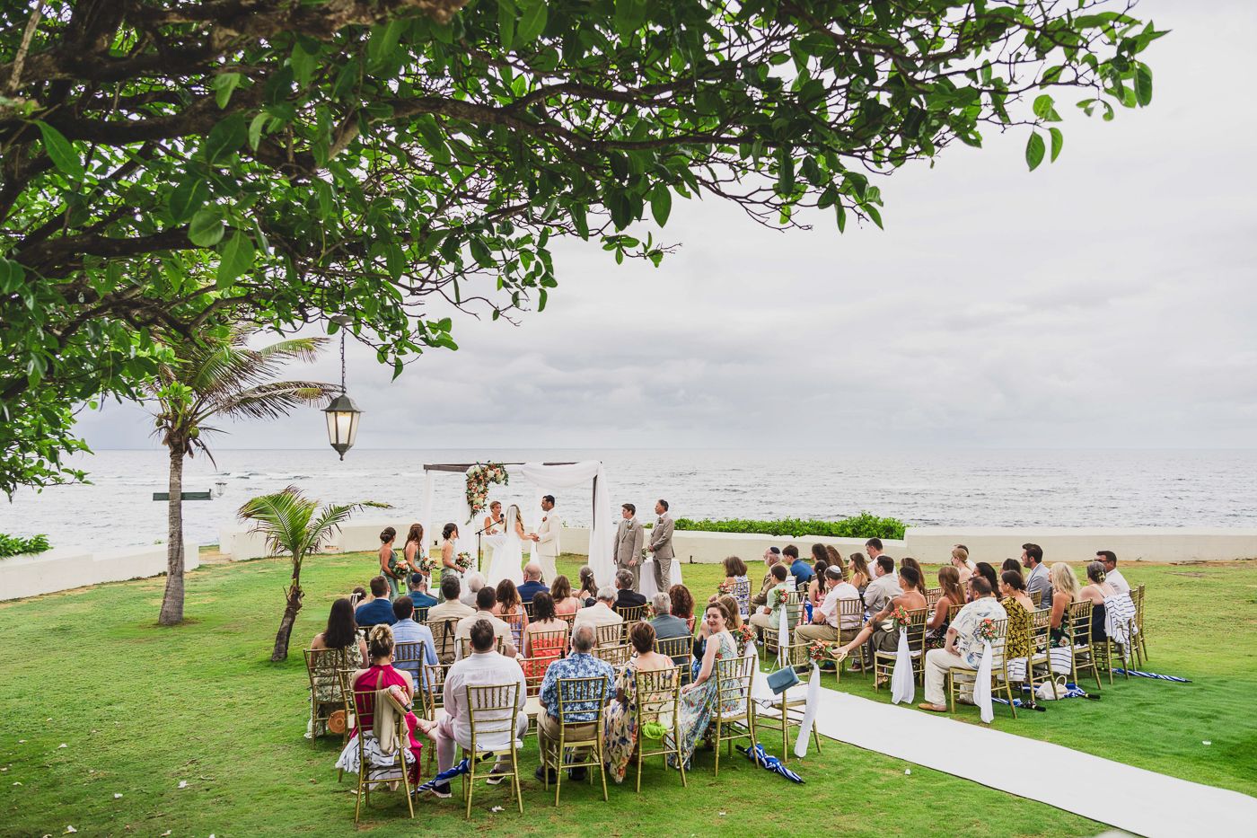 Outdoor beach wedding ceremony with guests seated on chairs facing couple at altar on green lawn by ocean.