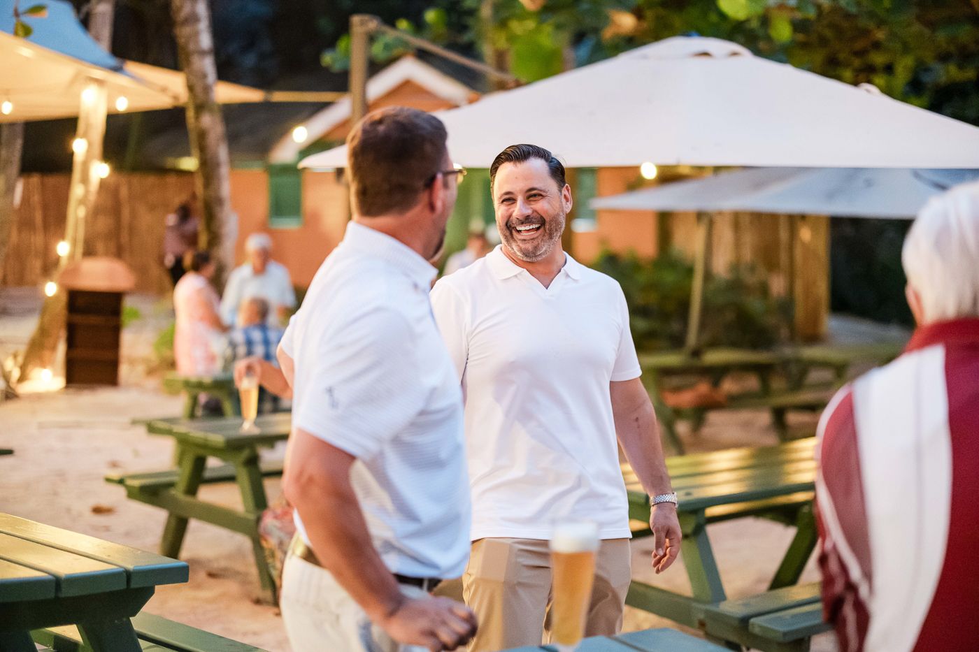 Two men in white shirts laughing and socializing at an outdoor beer garden with picnic tables.