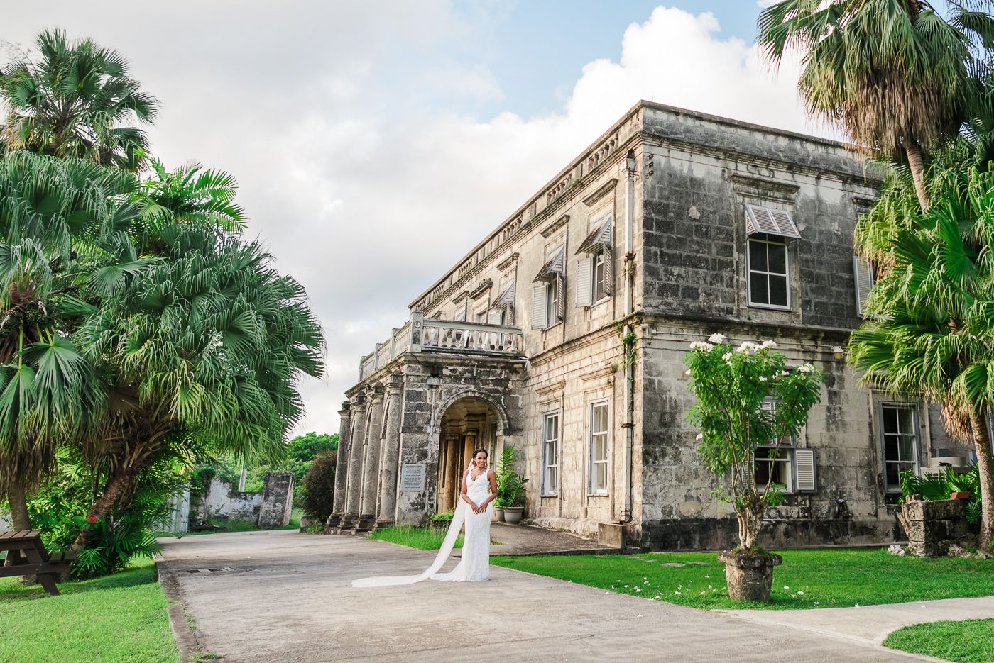 Bride in elegant white gown stands before a grand historic stone mansion with tropical gardens.