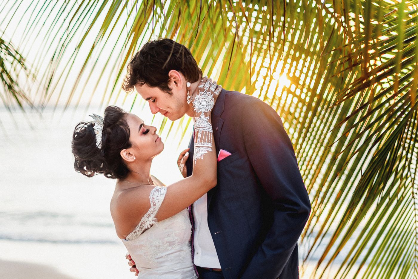 Bride and groom sharing a romantic moment under palm trees on a tropical beach at sunset.