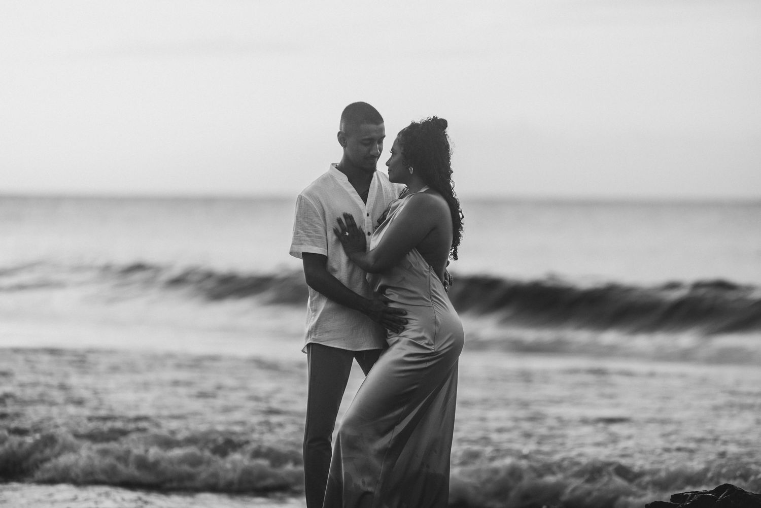 Black and white photo of a romantic couple embracing on a beach with ocean waves in the background.
