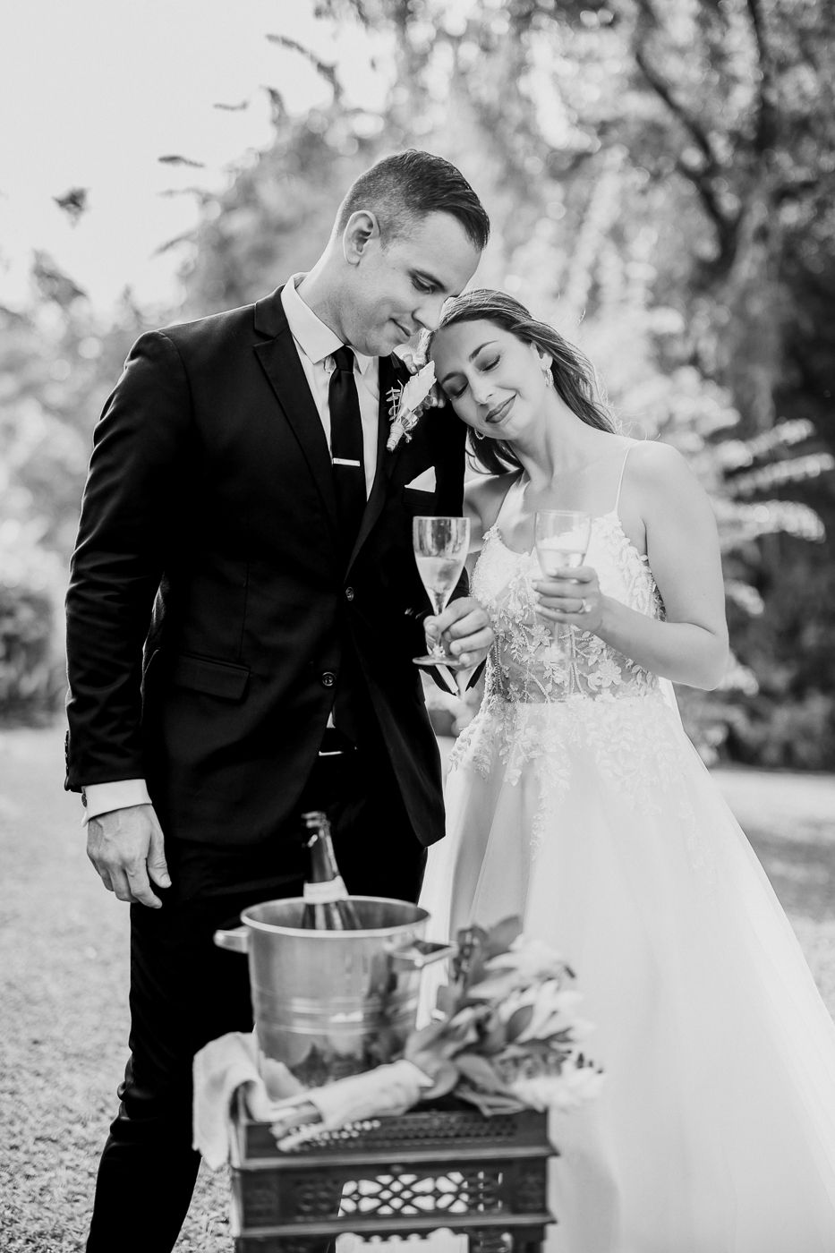 Black and white photo of a bride and groom holding champagne glasses beside an ice bucket at an outdoor wedding.