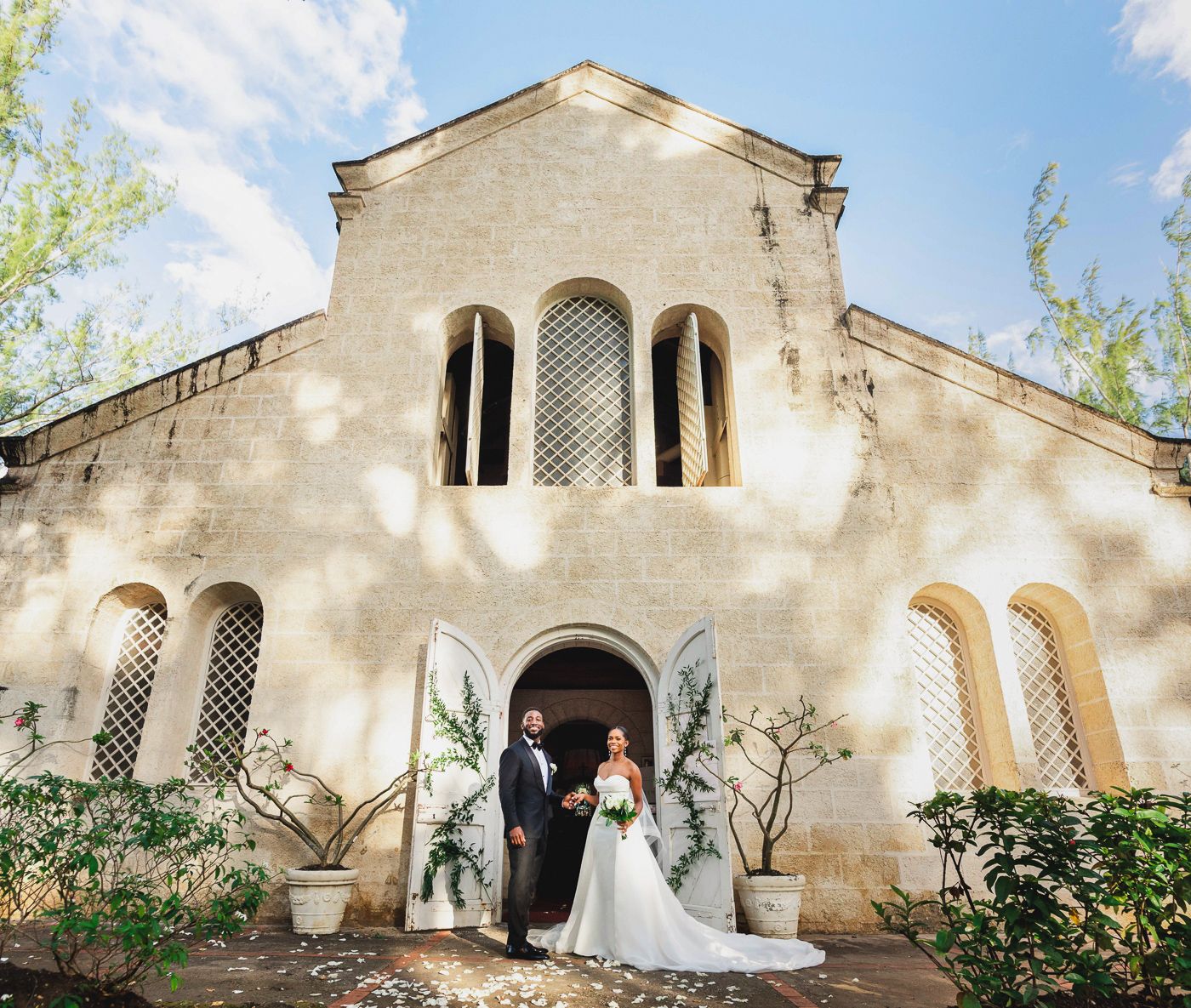 Bride and groom standing at the entrance of a beautiful stone chapel surrounded by lush greenery.