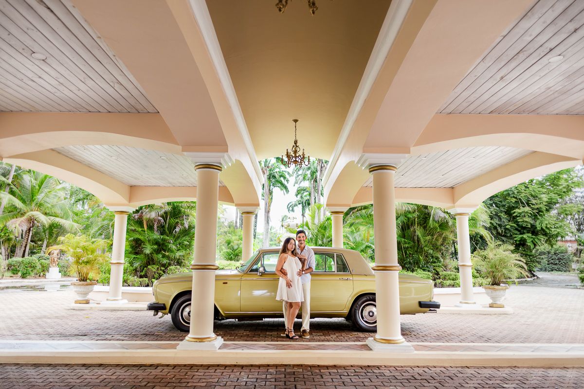 A vintage cream-colored car parked under an elegant covered driveway with tropical landscaping in the background.