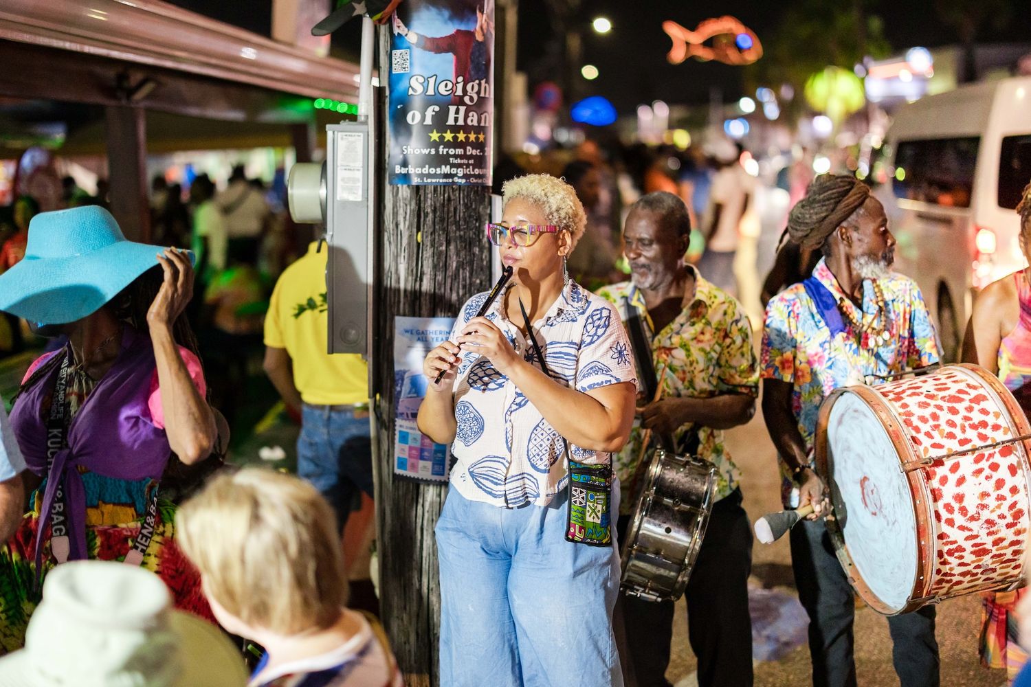 Colorful street musicians perform at a lively nighttime Caribbean festival, with a guitarist and drummer in vibrant attire.