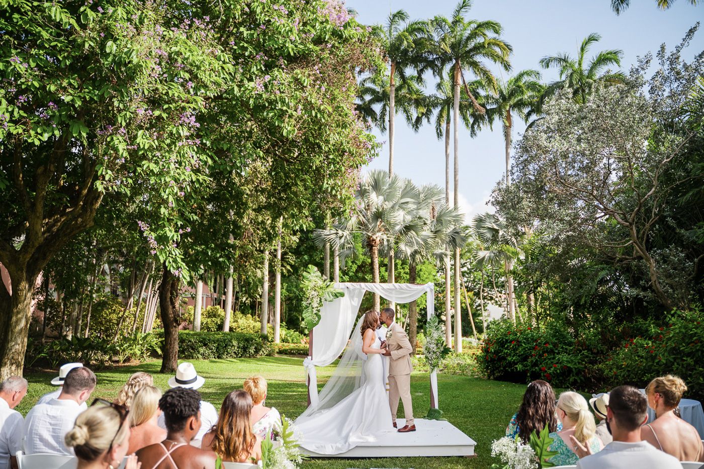 Garden wedding ceremony setup with white chairs and tropical landscaping in the background.