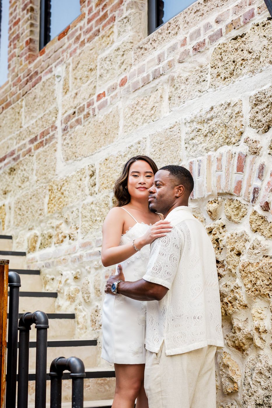 A romantic couple embrace on stone steps wearing matching white outfits on a sunny day. Sandy Lane barbados