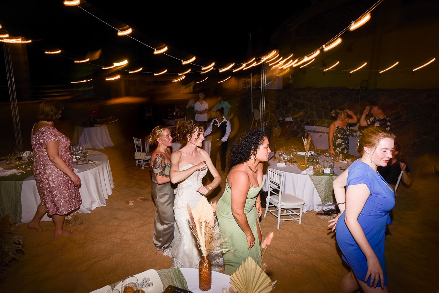 Wedding guests dance and celebrate on a sandy beach venue with overhead lighting.
