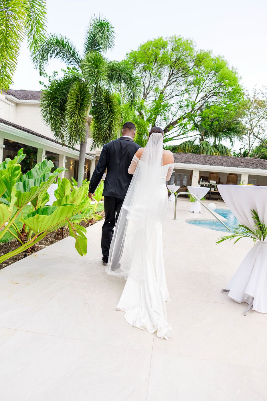 A couple walks along a white pathway with palm trees and a pool in the background.