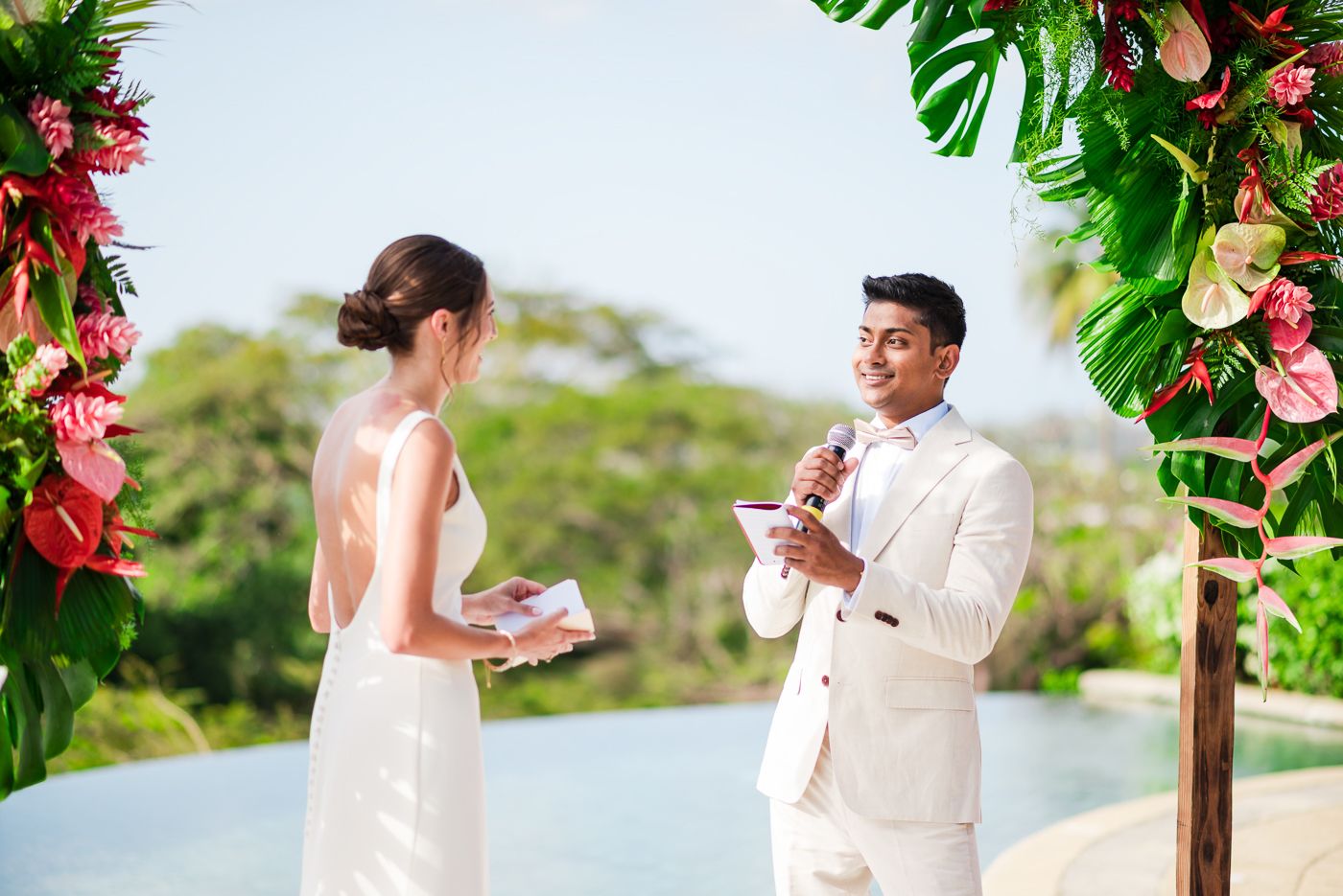 Couple sharing vows under tropical arch with infinity pool view.