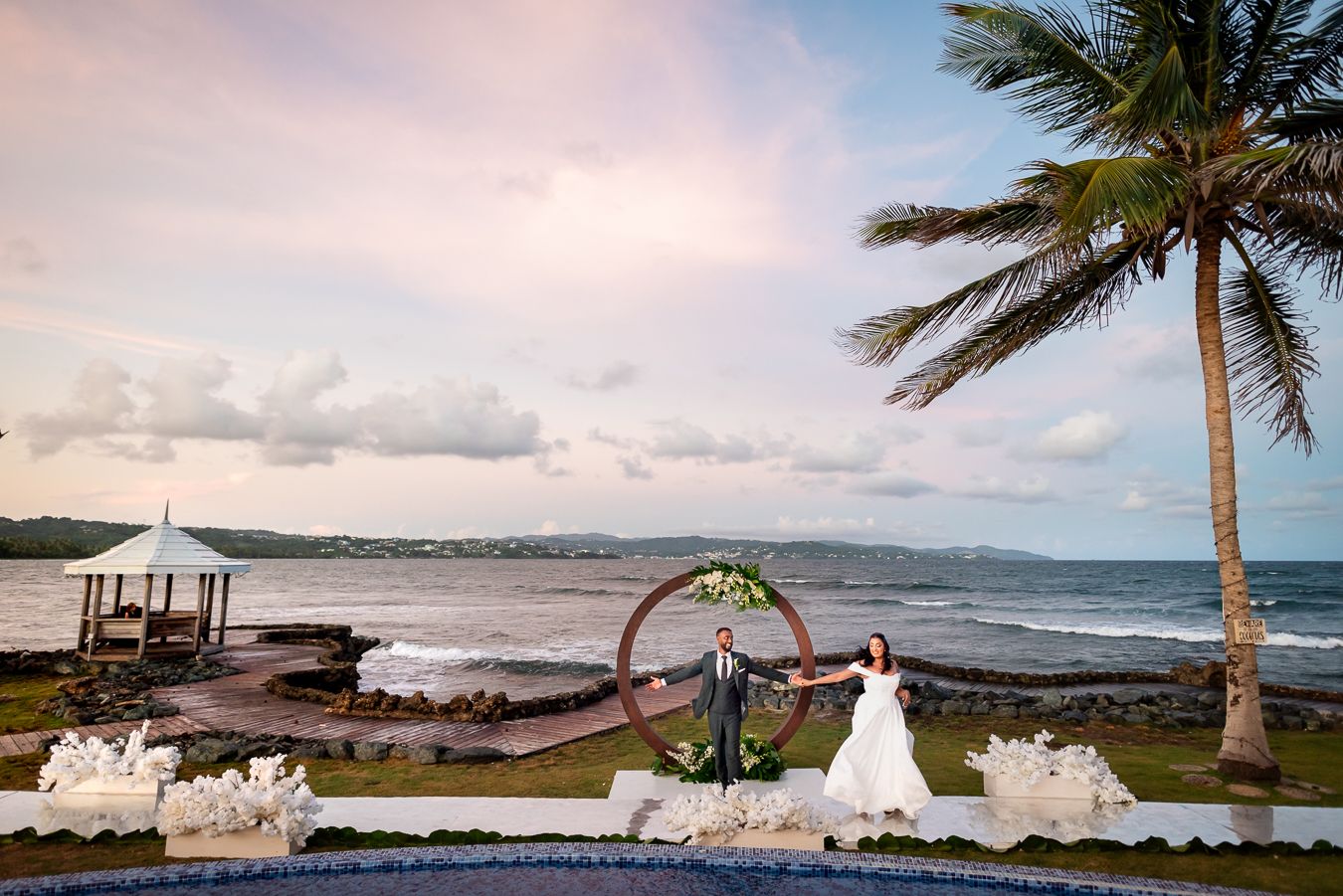 Scenic oceanfront wedding ceremony setup with palm trees and white floral arrangements.