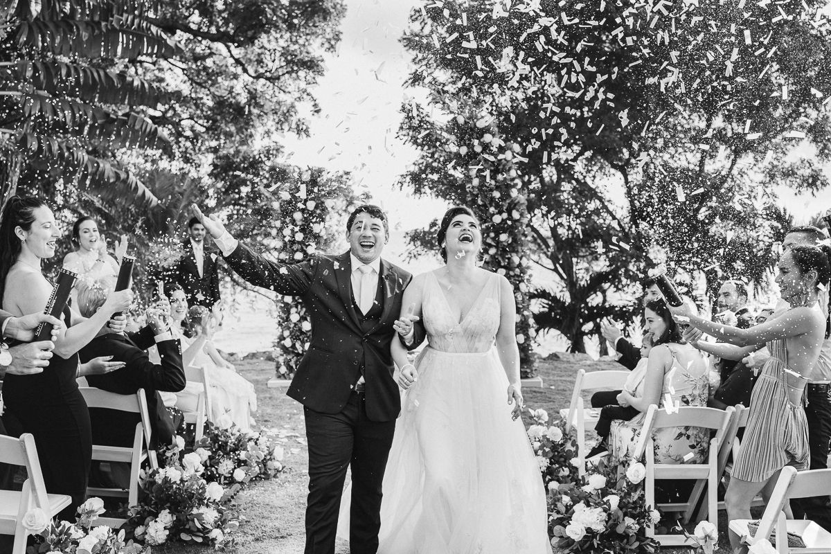 Black and white photo of a joyful bride and groom walking down the aisle as guests toss flower petals.