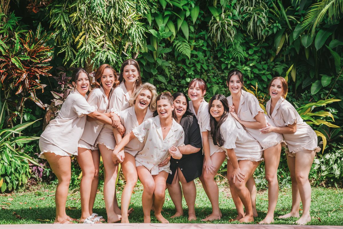 A group in matching blush pink robes poses together outdoors against lush tropical greenery in a garden setting.
