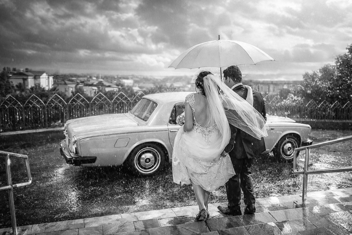 Dramatic black and white panorama of vintage car and wedding couple in rainy weather.