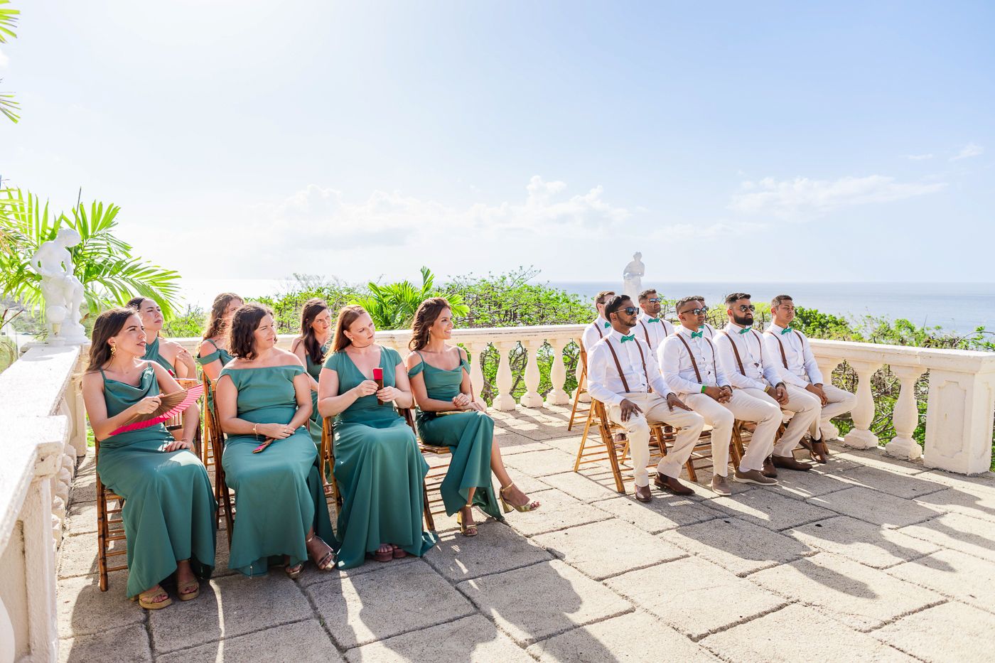 Wedding party in green dresses and khaki suits seated on stone terrace overlooking scenery.