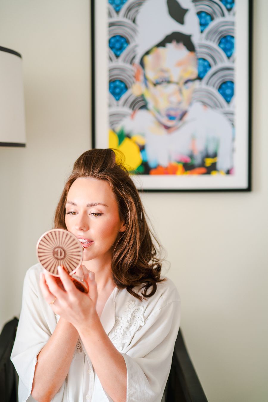 A person in white holds a small pink handheld fan while sitting in front of colorful wall art.
