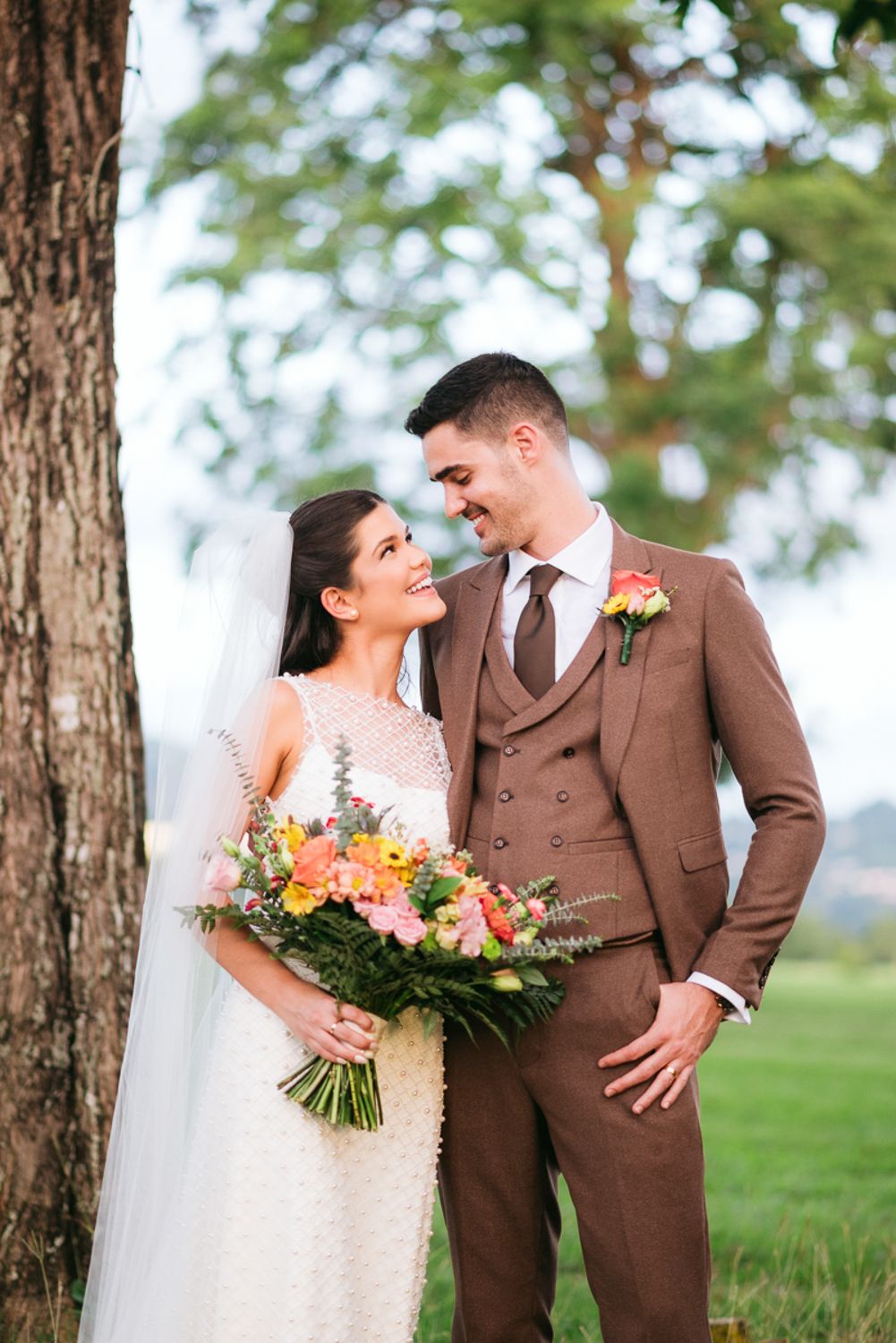 A bride in a white lace dress holds a peach and pink bouquet next to her groom wearing a brown suit outdoors.