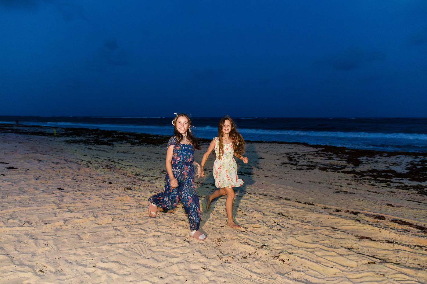 Two girls holding hands and walking on a sandy beach at dusk with ocean waves in the background.