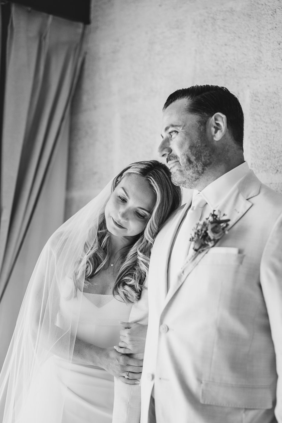 Black and white photo of a bride leaning on groom's shoulder, both in wedding attire, sharing a tender moment.
