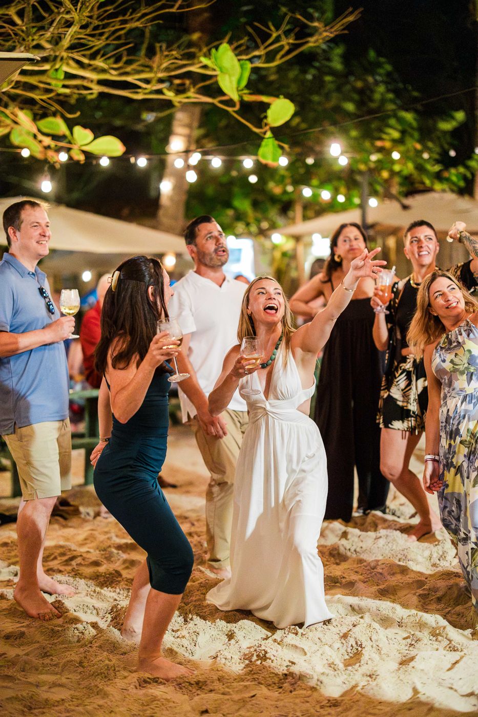 Bride in white dress laughing with guests at an outdoor beach wedding reception under string lights at night.
