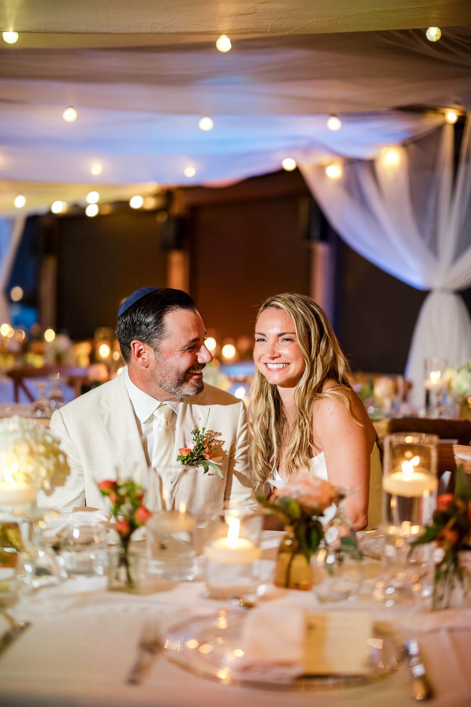 Bride and groom share a joyful moment at their candlelit wedding reception table decorated with floral centerpieces.