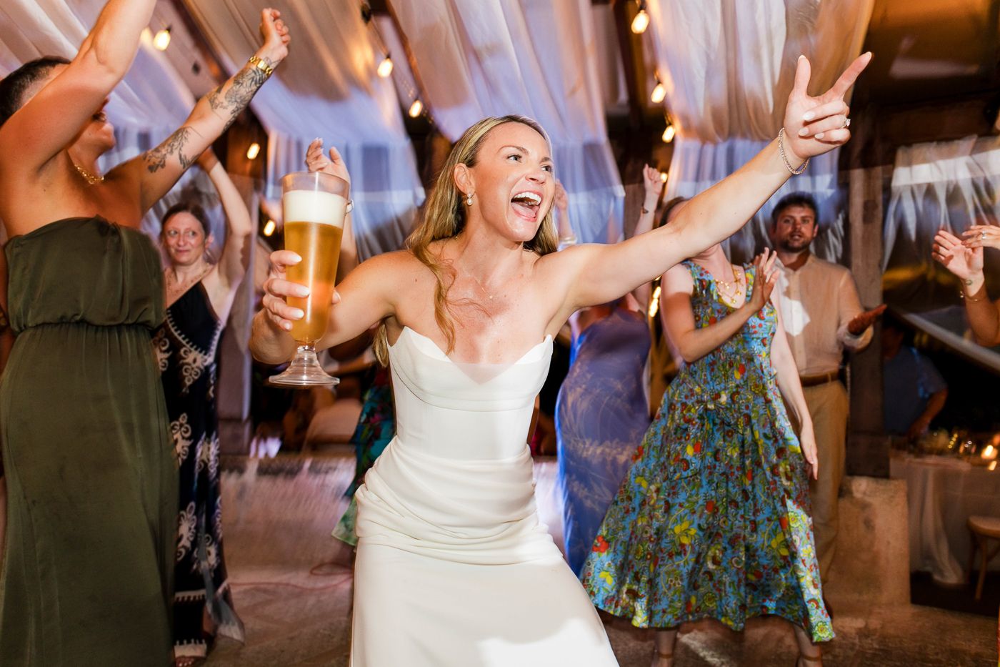Joyful bride in white gown holding beer glass with arms raised at wedding reception celebration.