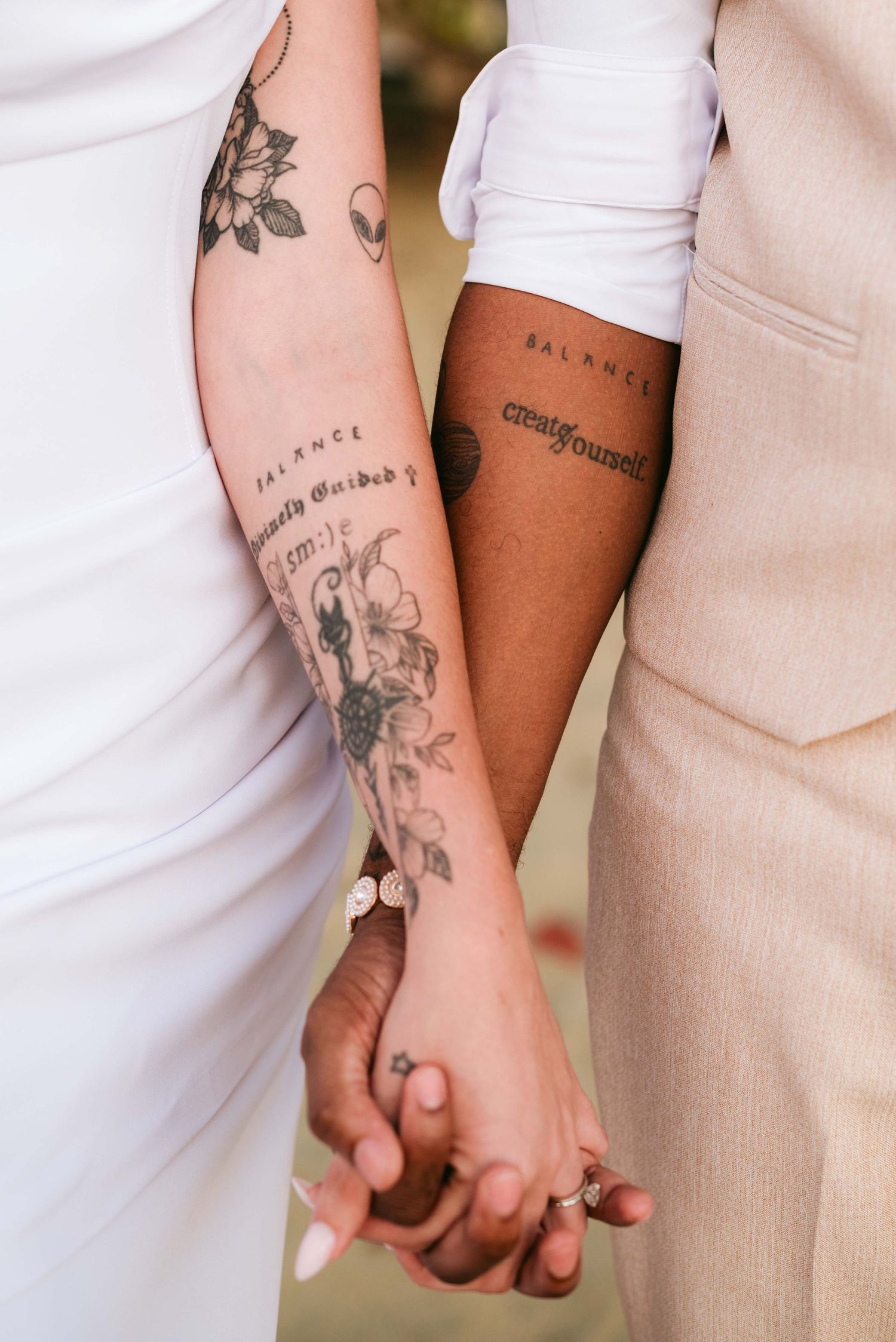 Close-up of tattooed arms holding hands against a light background, showing multiple floral and text tattoo designs.