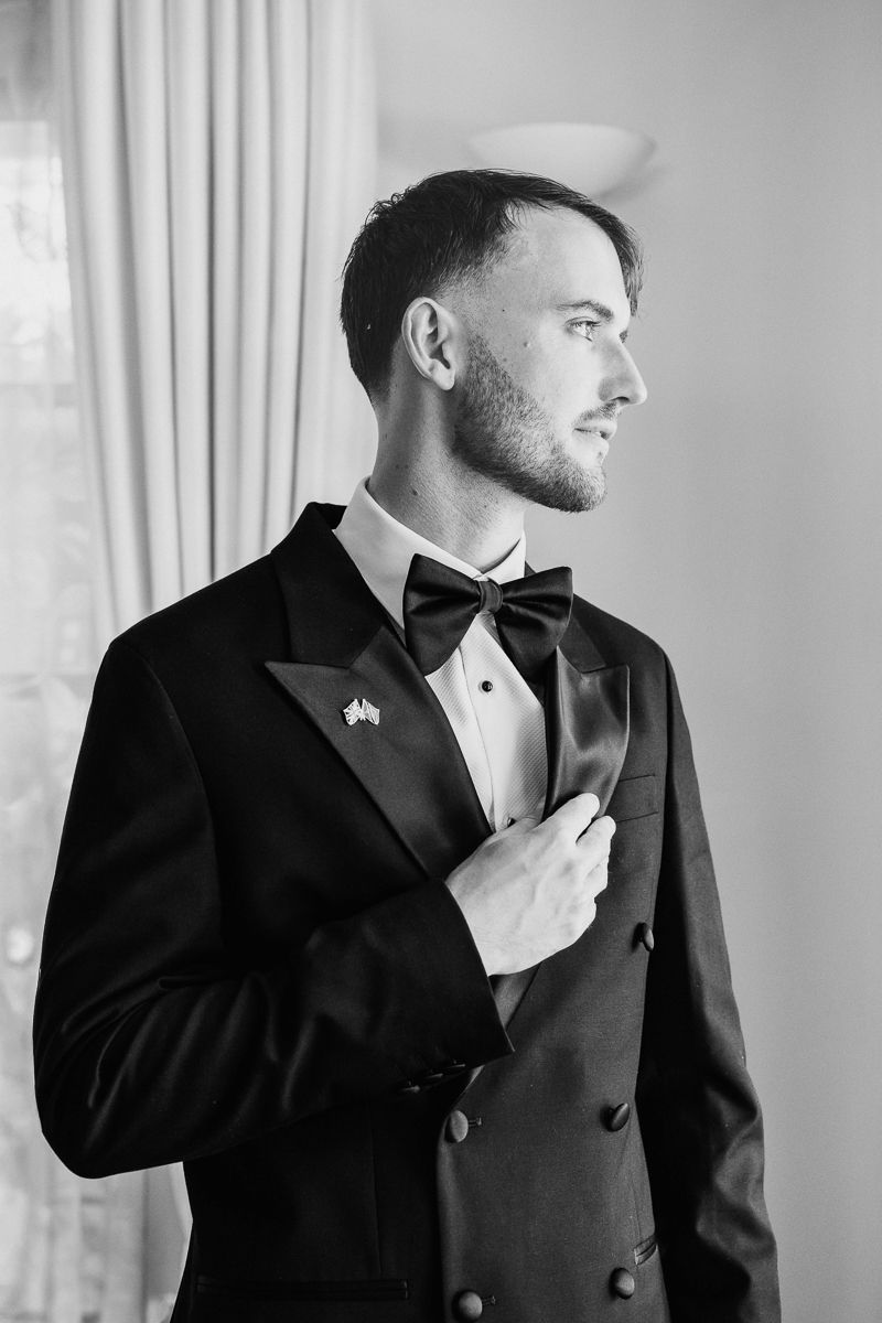 A man in a black tuxedo adjusts his bowtie while looking to the side in a formal black and white portrait.