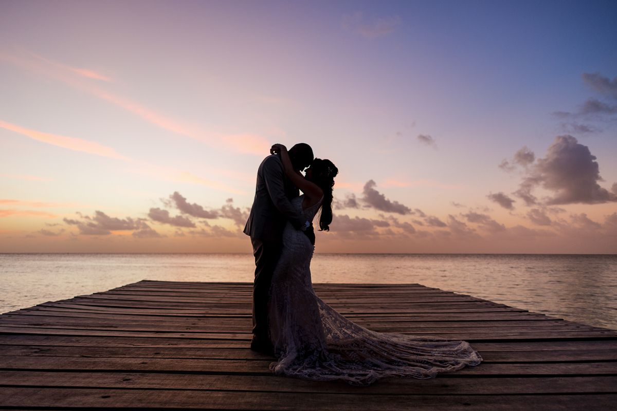 Silhouette of a bride and groom kissing on a wooden pier at sunset over calm ocean waters.