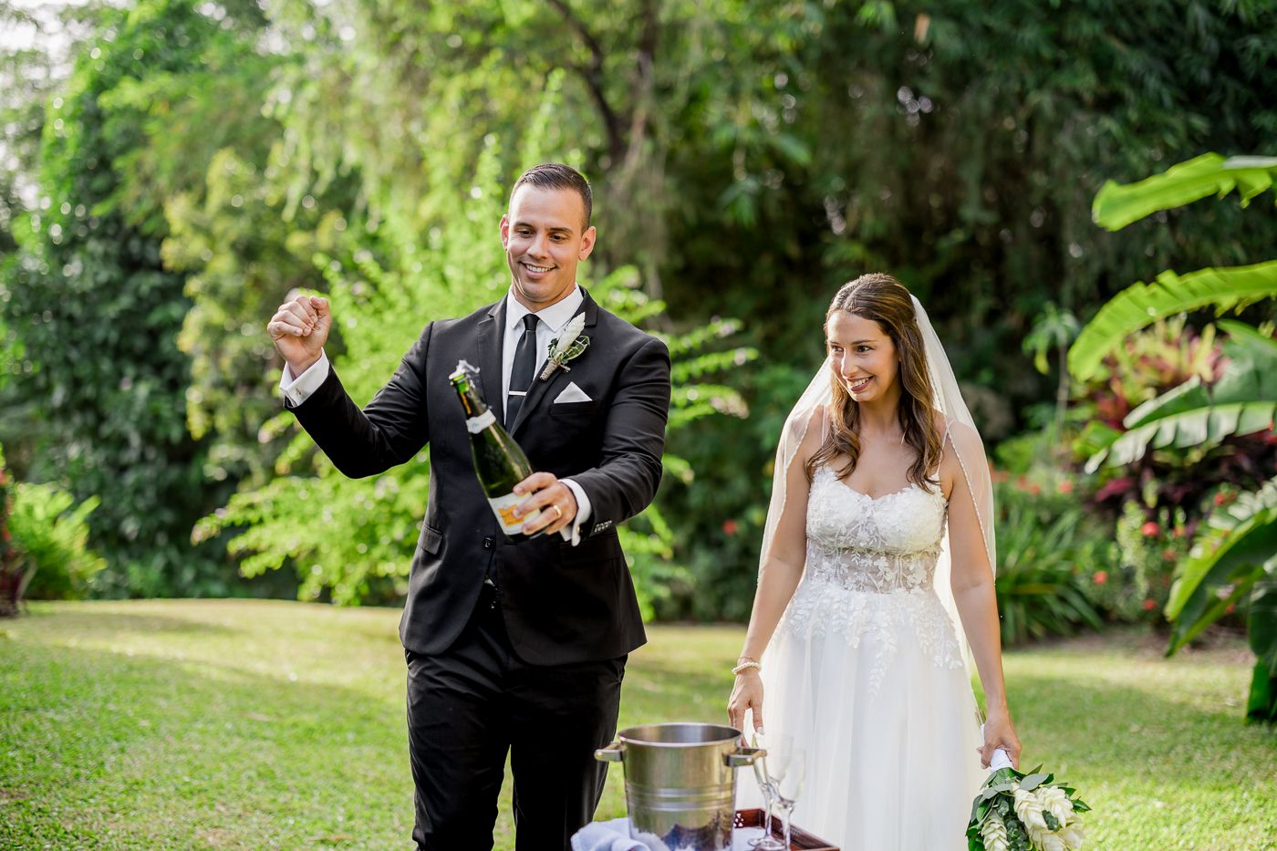 Bride and groom celebrating outdoors, groom opening champagne bottle while bride holds bouquet in lush green garden.