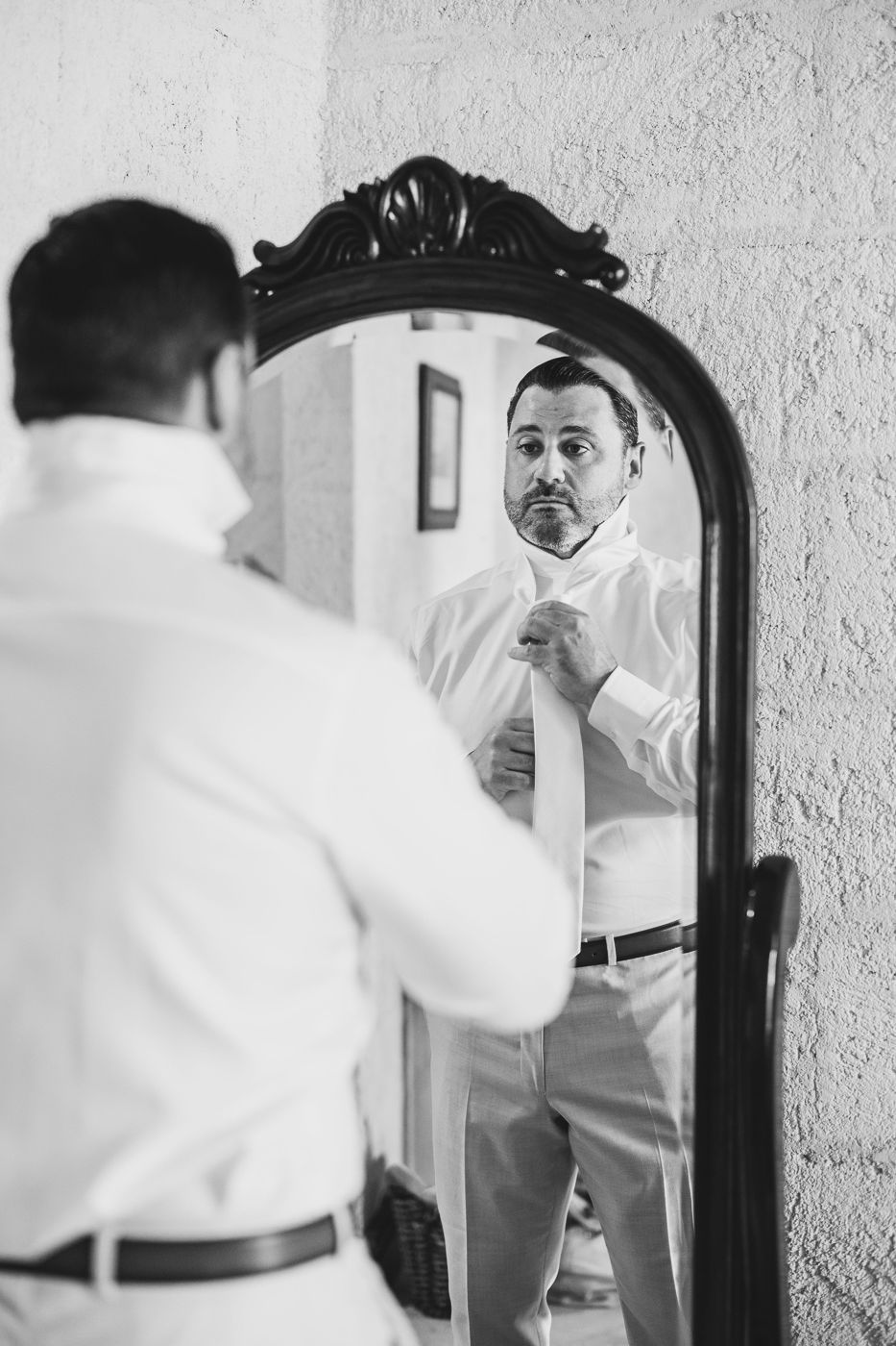 Black and white photo of a groom adjusting his tie while looking at his reflection in an ornate floor mirror.