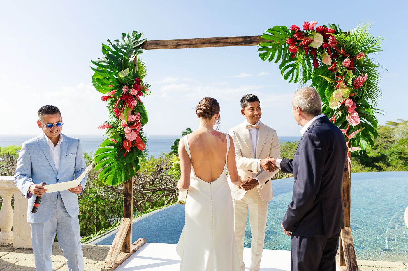 Wedding ceremony takes place under wooden arch with tropical flowers and ocean backdrop.