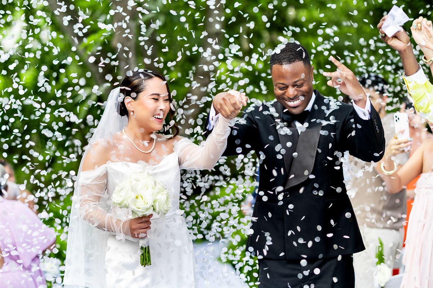 Newlyweds celebrate with raised arms as confetti falls during their outdoor garden ceremony.