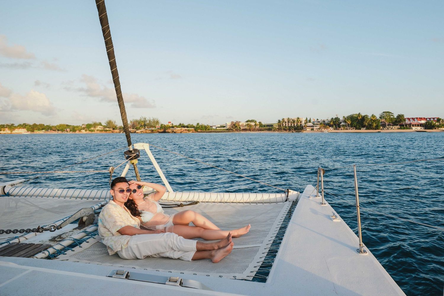 Couple relaxes on cushions at the bow of a catamaran sailboat while cruising tropical waters at sunset.